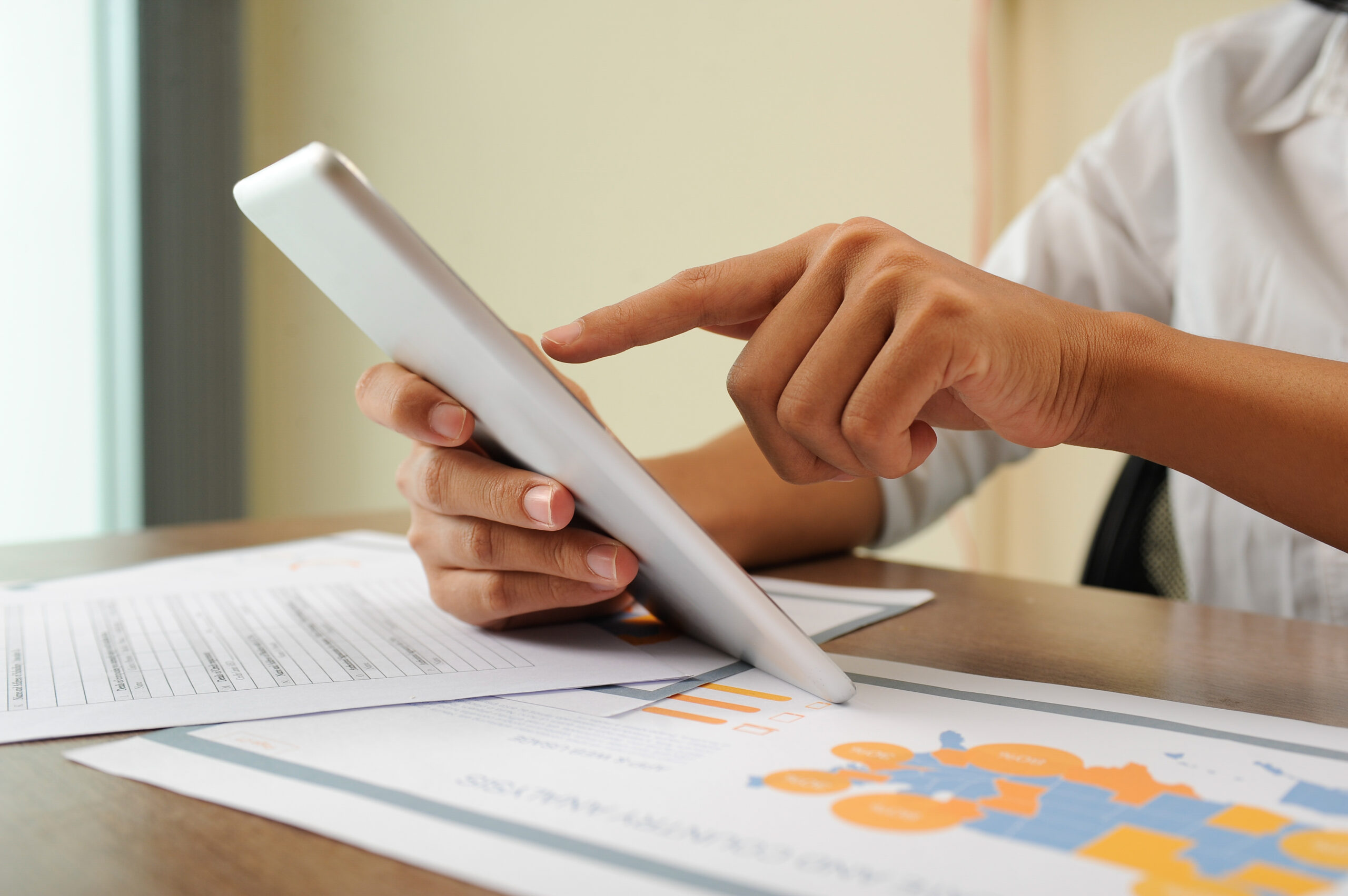 Closeup of person holding and using tablet. Papers lying on desk. Marketing research concept. Cropped view.